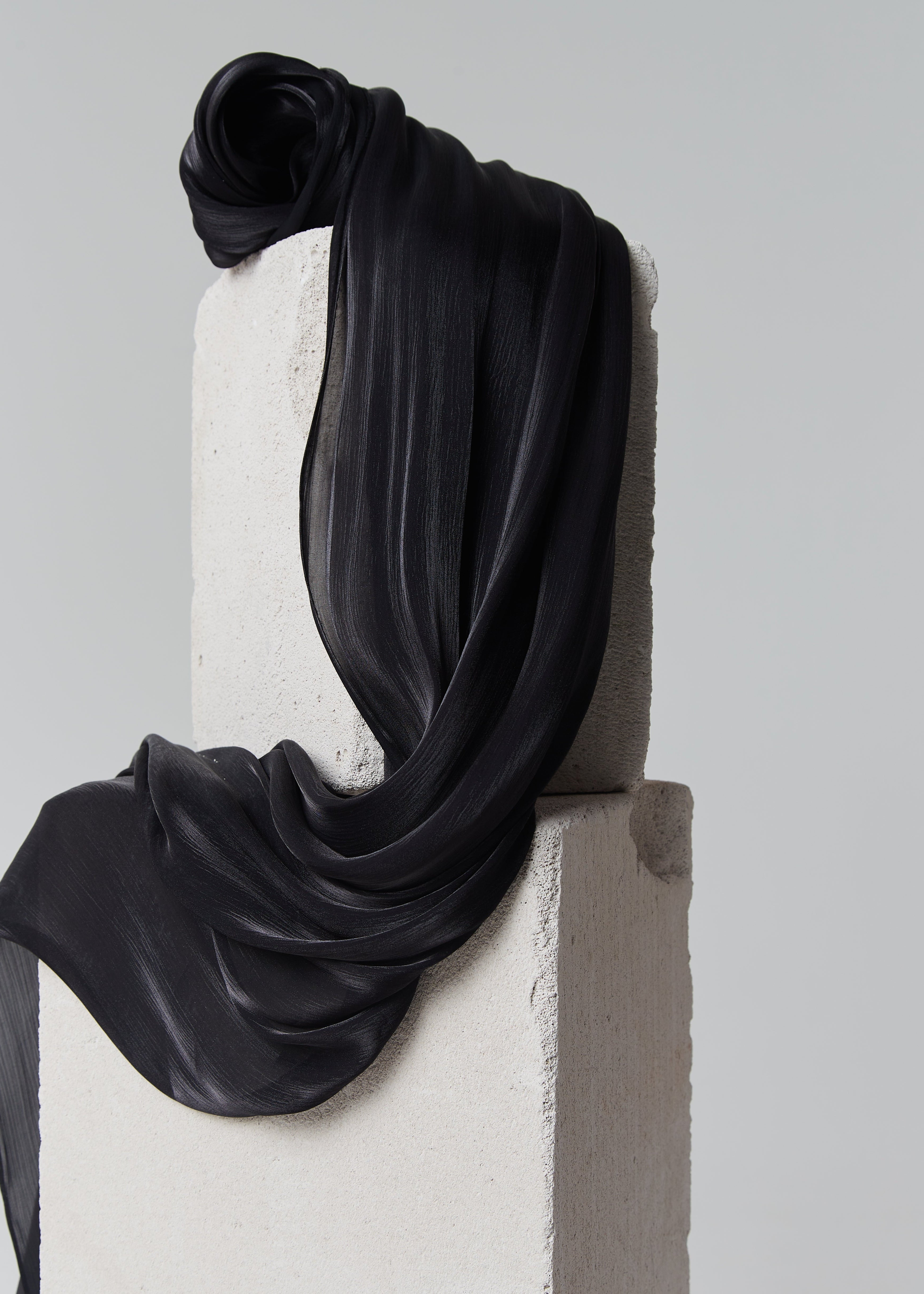 Close-up of black silk hijab from Yovlaq draped over concrete blocks – rich texture and matte-shine finish in a classic elegant tone.