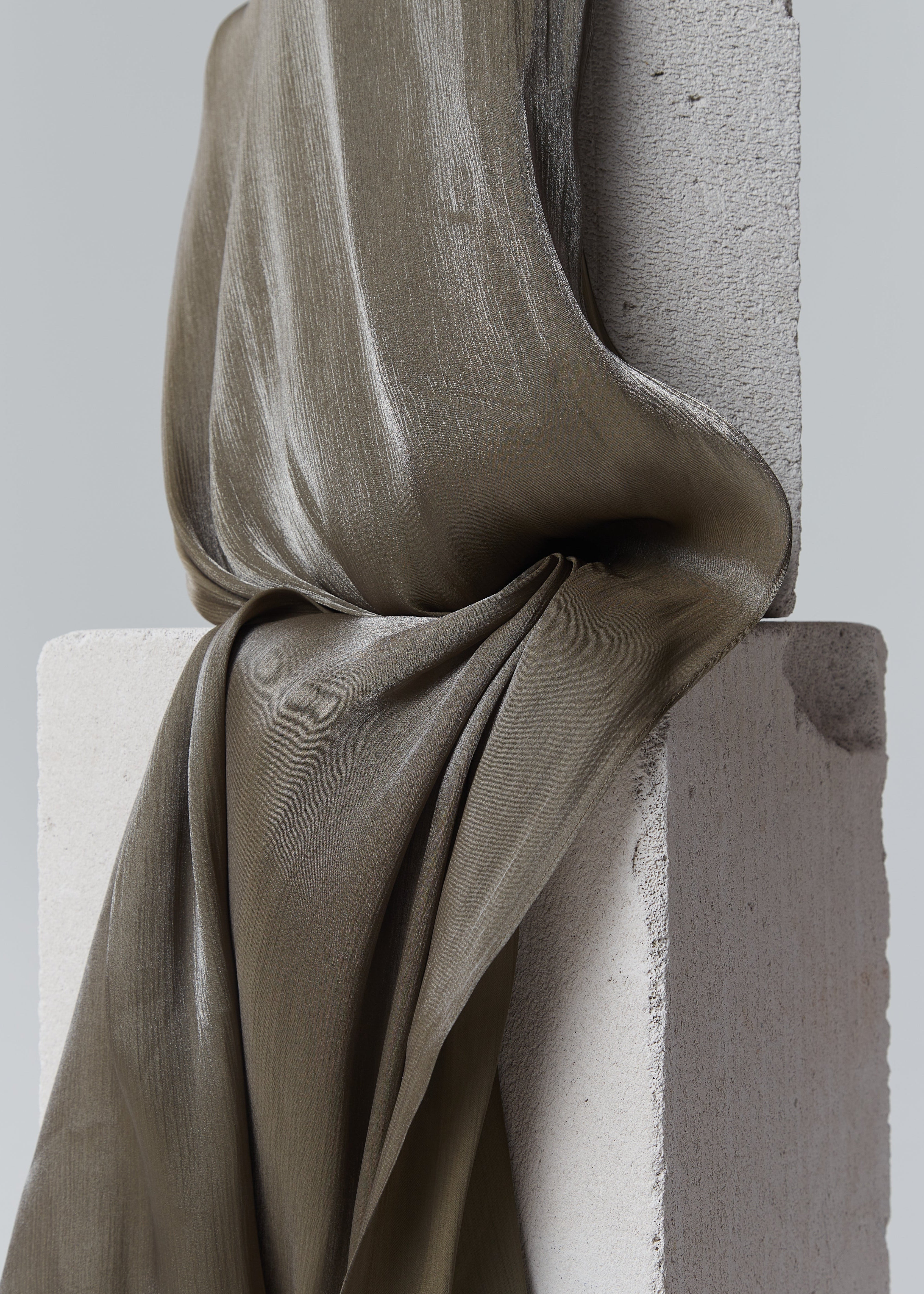 Khaki ready-to-wear silk hijab draped over minimal concrete blocks, showcasing its rich sheen and elegant texture.