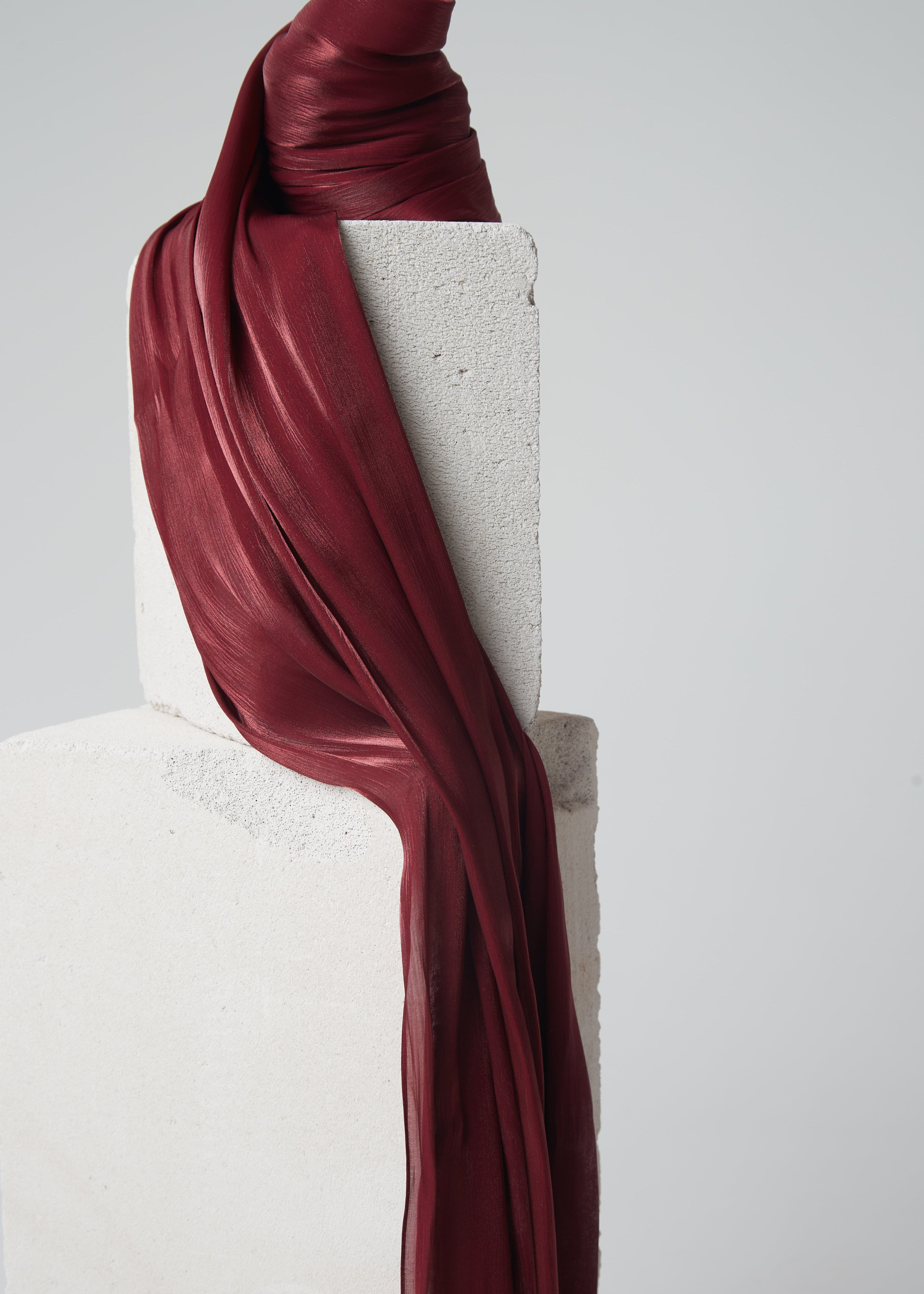 Ready-to-wear silk hijab in a rich maroon shade, elegantly draped over minimalist stone blocks, showcasing its glossy texture and deep colour.