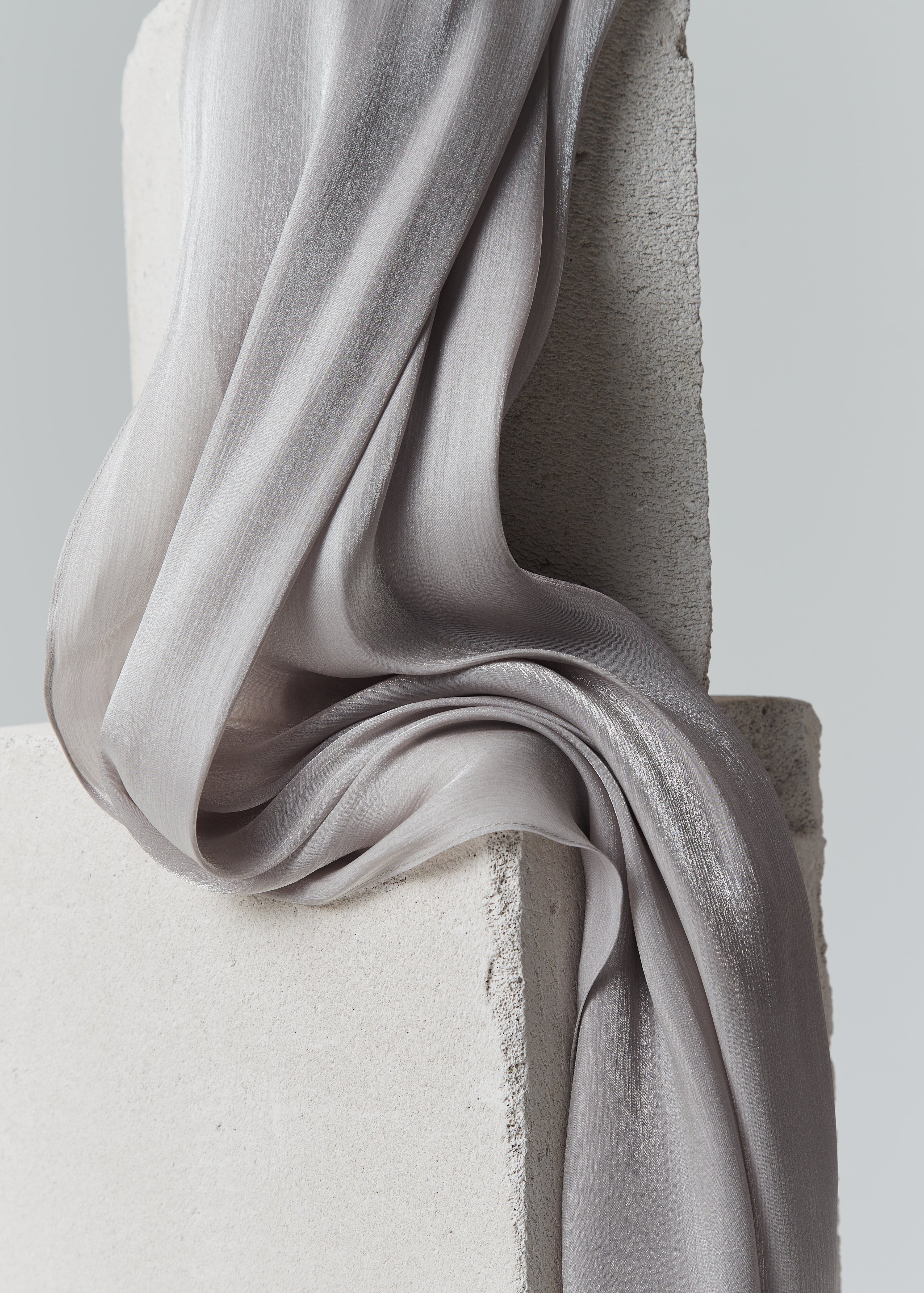 Close-up of taupe grey silk hijab from Yovlaq draped over concrete blocks – flowing texture and soft matte-sheen finish.
