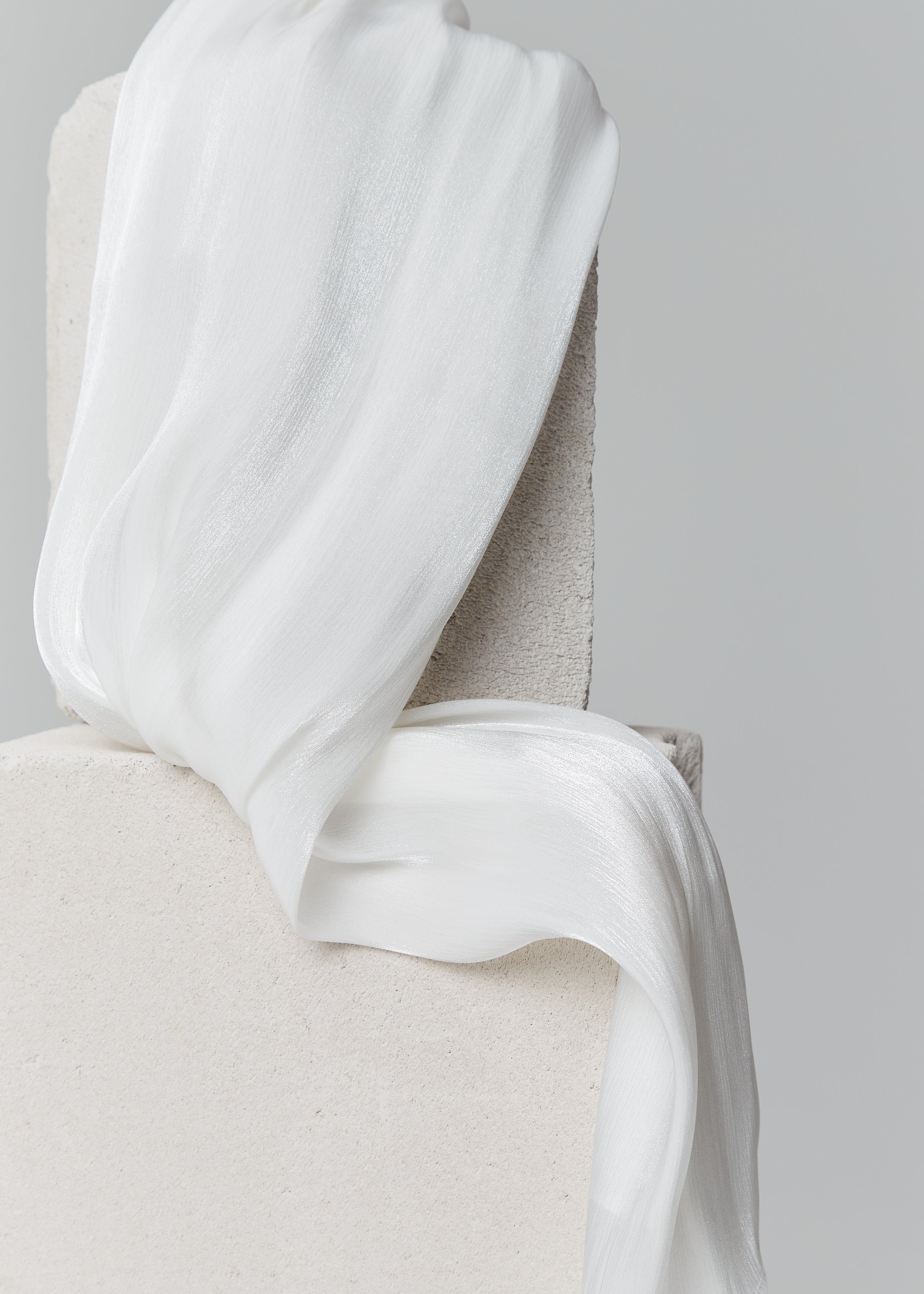 Glossy white ready-to-wear silk hijab elegantly draped over stacked stone blocks, highlighting its airy texture and soft shimmer.