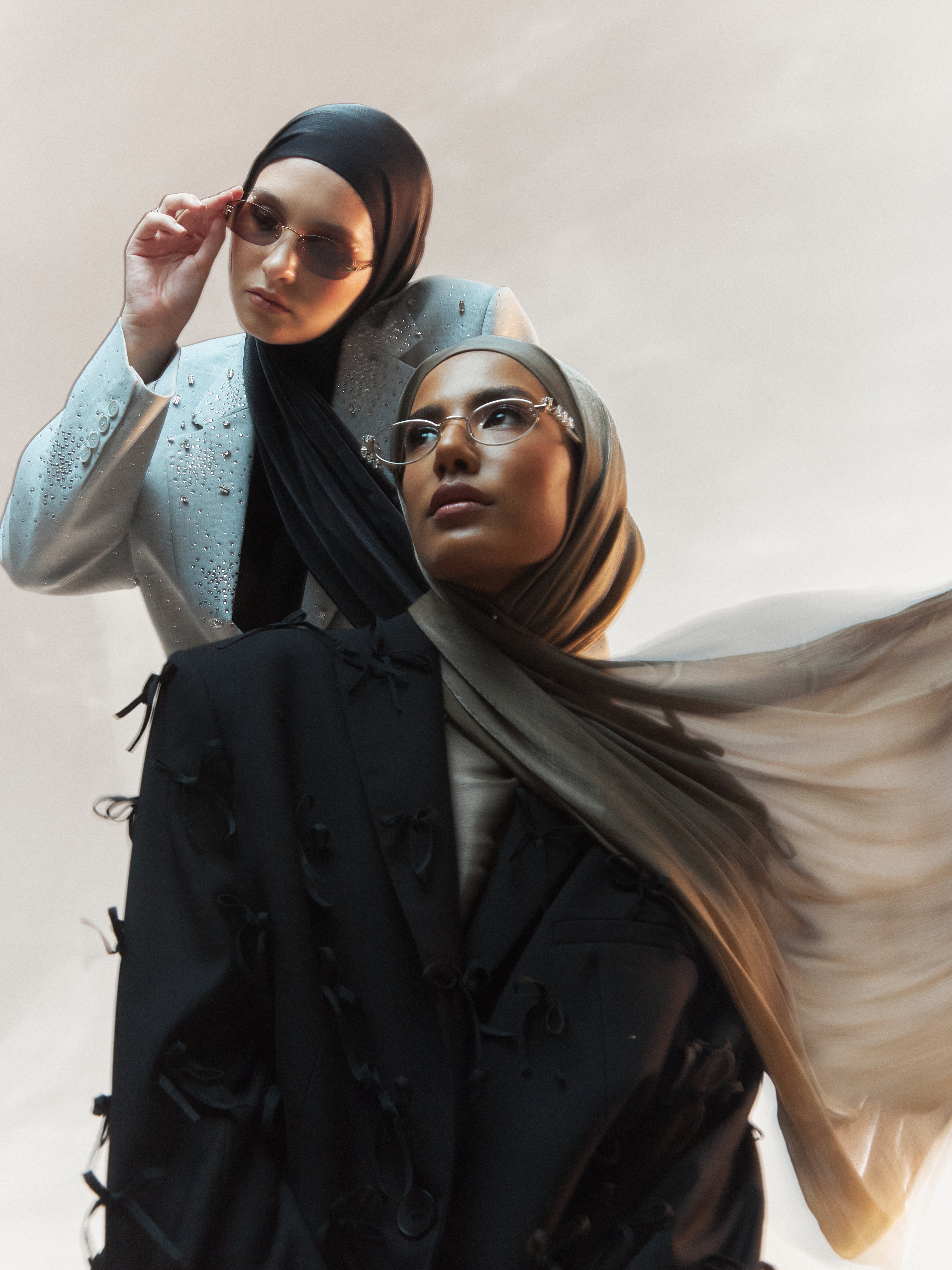 Model wearing a khaki ready-to-wear silk hijab styled with a black statement blazer, standing in front of another model in a black hijab and light blue embellished suit, both exuding confidence in an editorial fashion pose.
