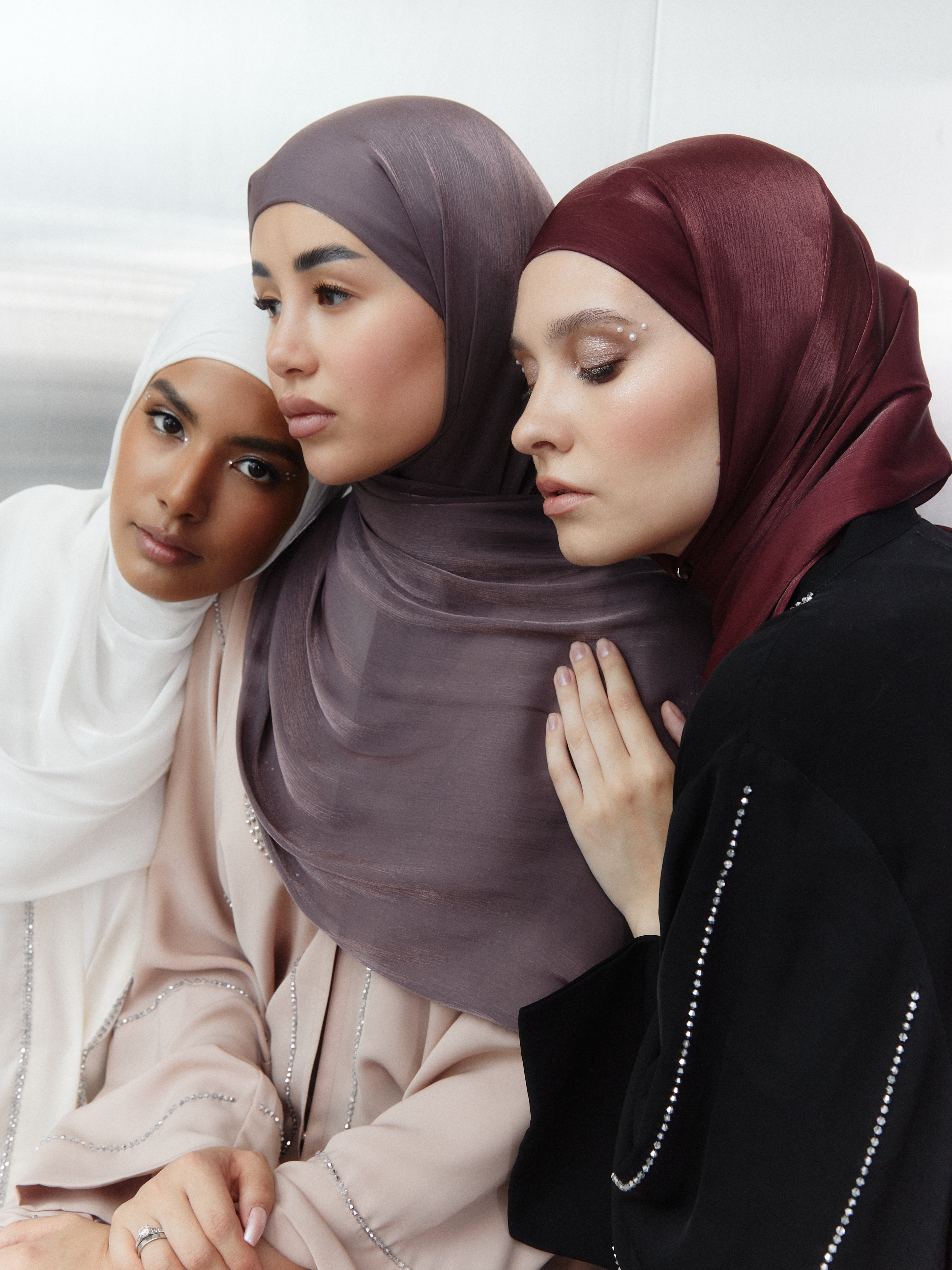 Three women wearing silk hijabs: in white, mauve, and deep red tones — posed close together with soft makeup and embellished outfits, showcasing colour contrast and texture in a fashion editorial style.