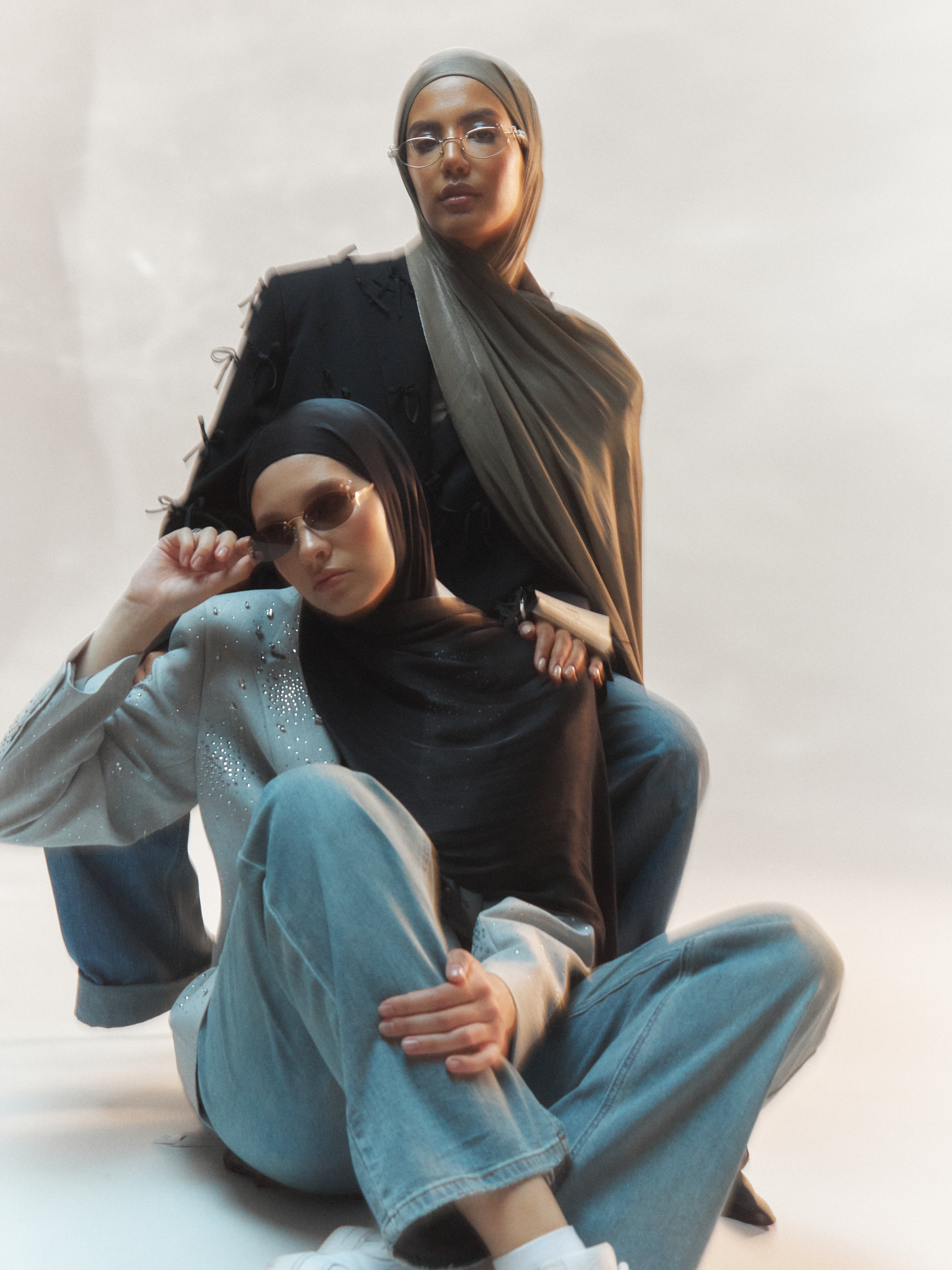Two women in Yovlaq’s black and khaki silk hijabs, styled with embellished jackets and denim, posing in a studio editorial with vintage sunglasses – expressive and modern modest fashion.