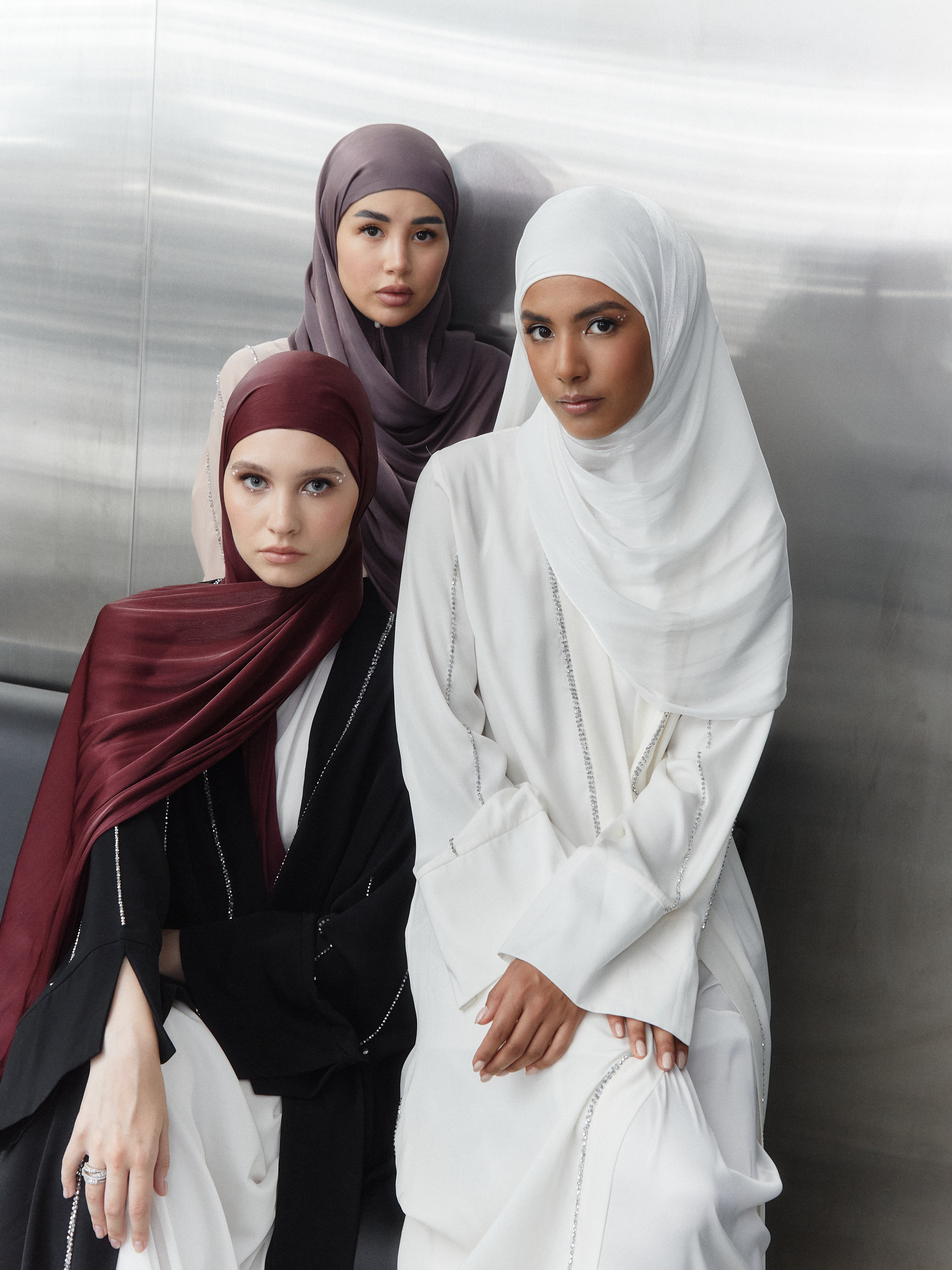 Three women modeling silk hijabs in deep red, crisp white, and soft mauve shades — styled with embellished modestwear in a modern, polished setting.