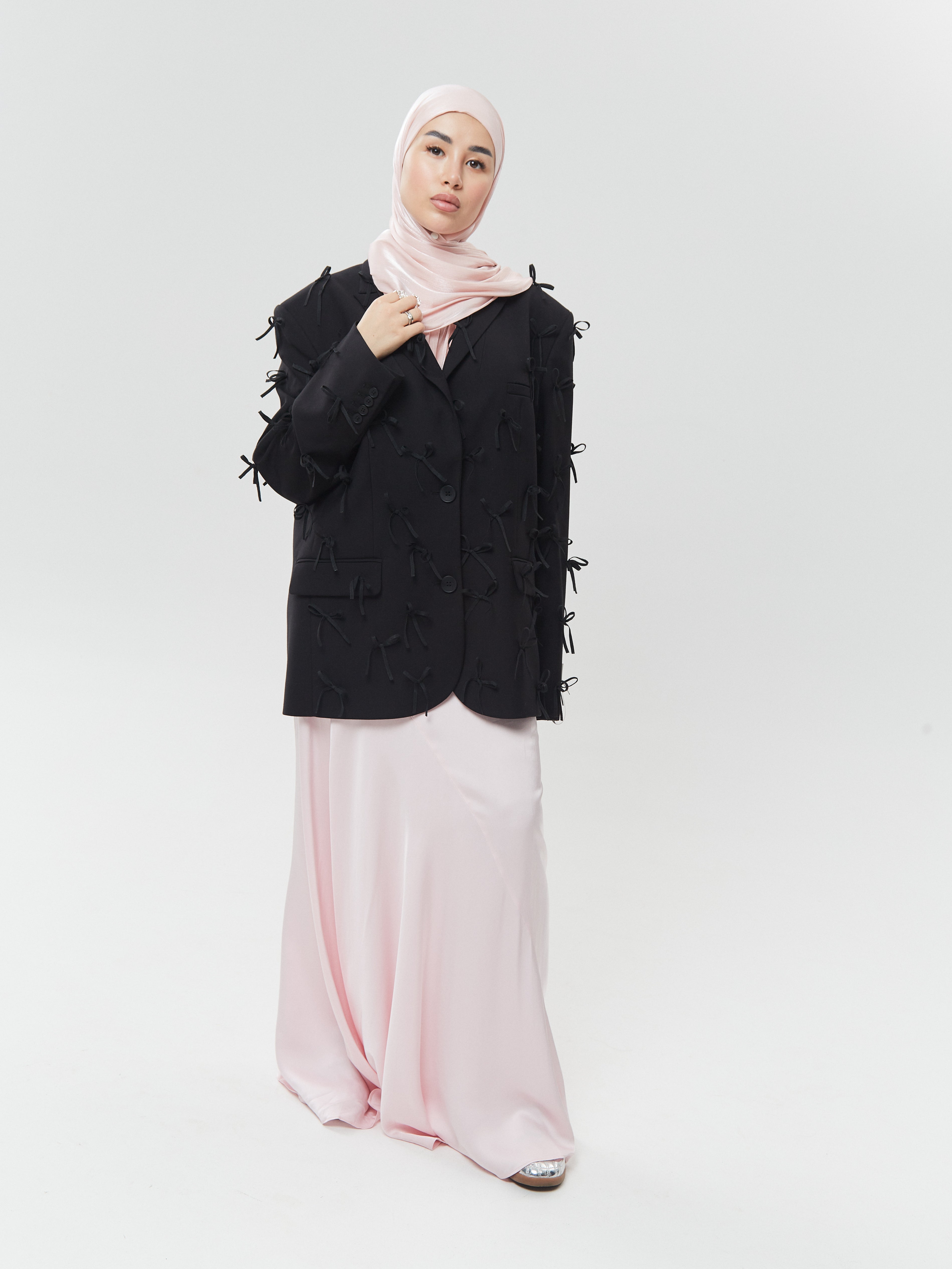 Full-length view of woman in pastel pink silk hijab from Yovlaq, styled with a bow-detailed black blazer and flowing blush satin skirt – elevated modern modest fashion.