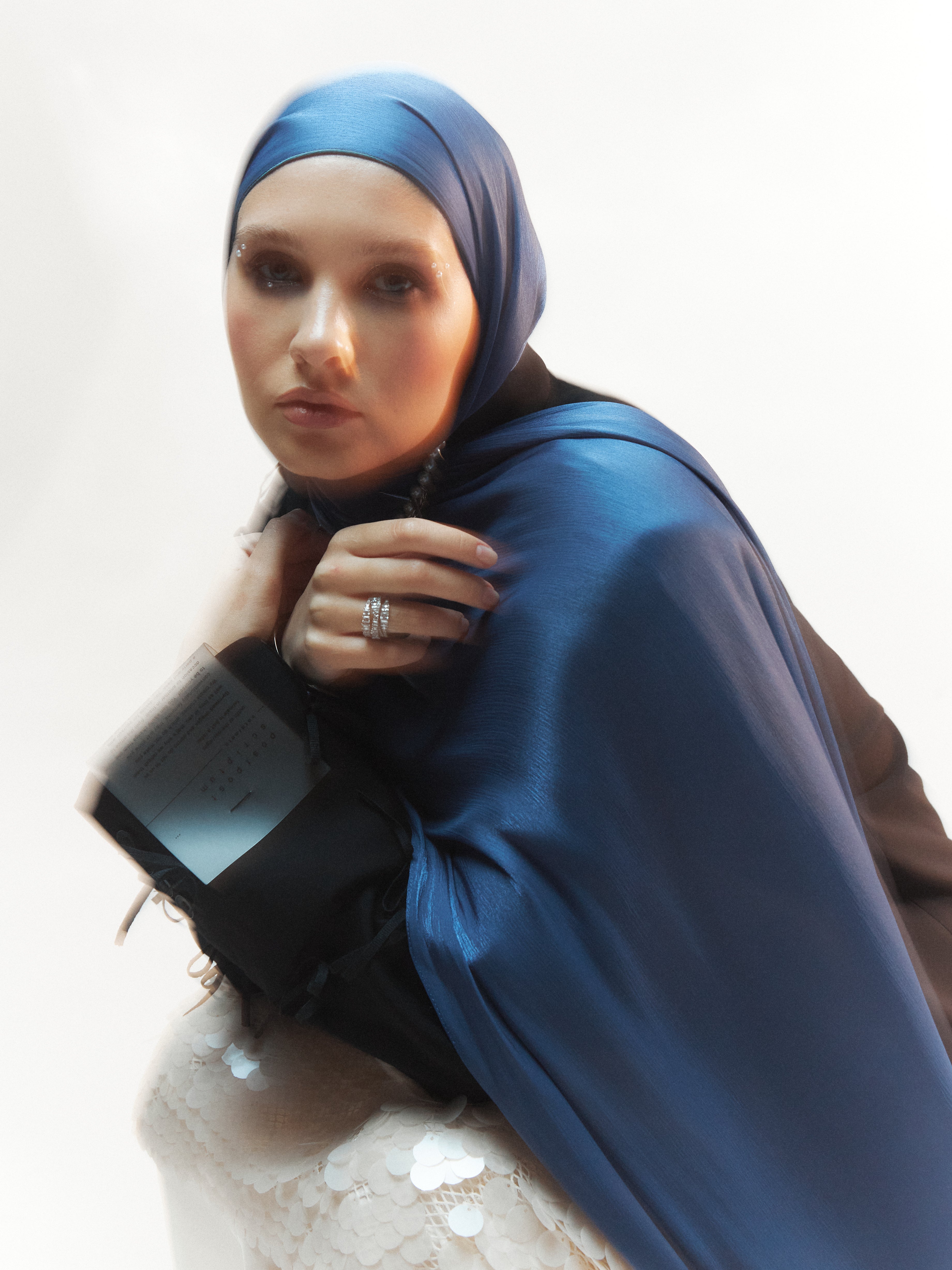 Close-up of a woman wearing a steel blue ready-to-wear silk hijab paired with a black blazer and white sequin skirt, seated against a softly blurred background.