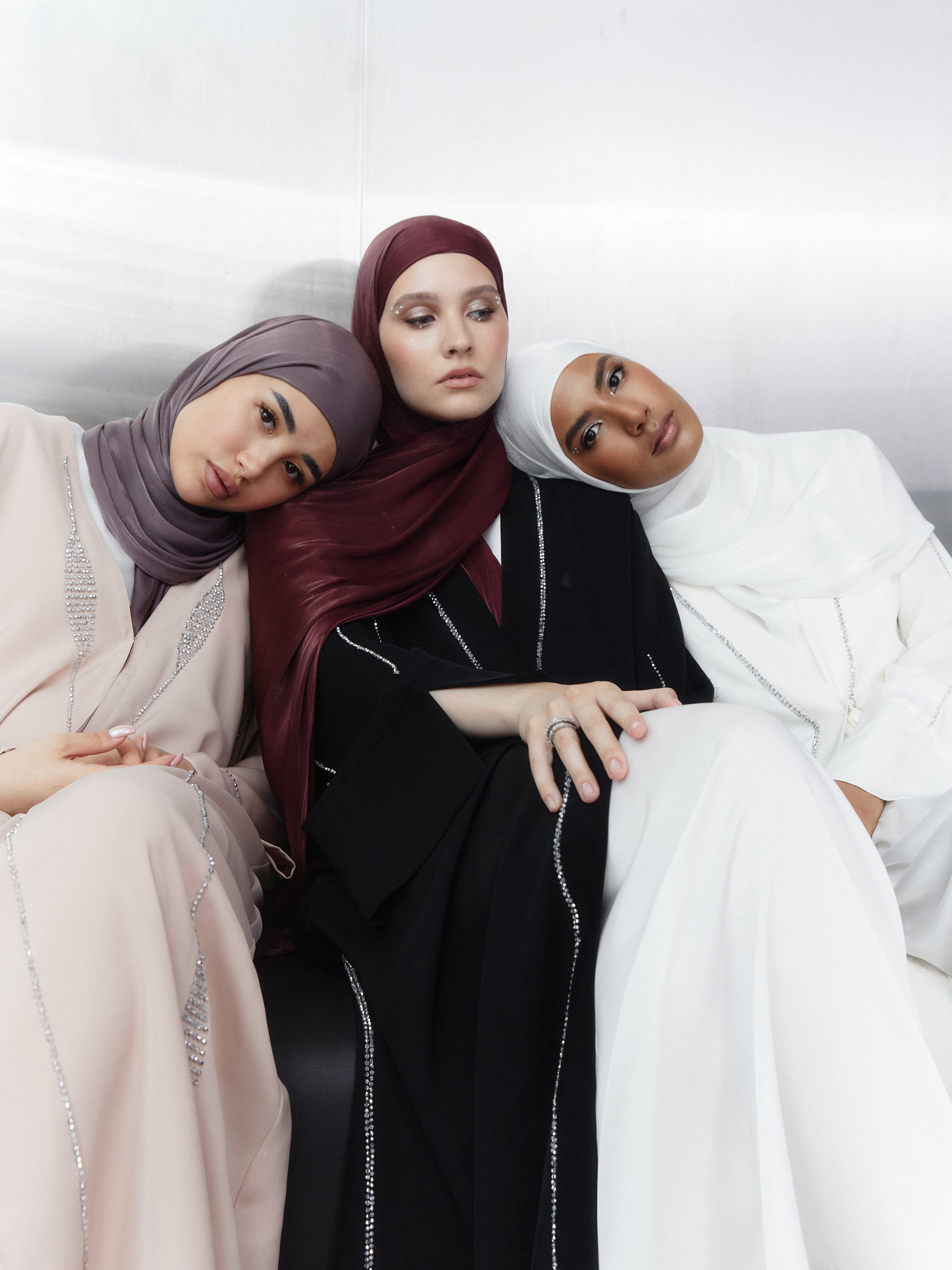 Three women resting together in soft-toned hijabs — featuring shades of mauve, red, and ivory white, styled with flowing embellished abayas in a serene, contemporary scene.
