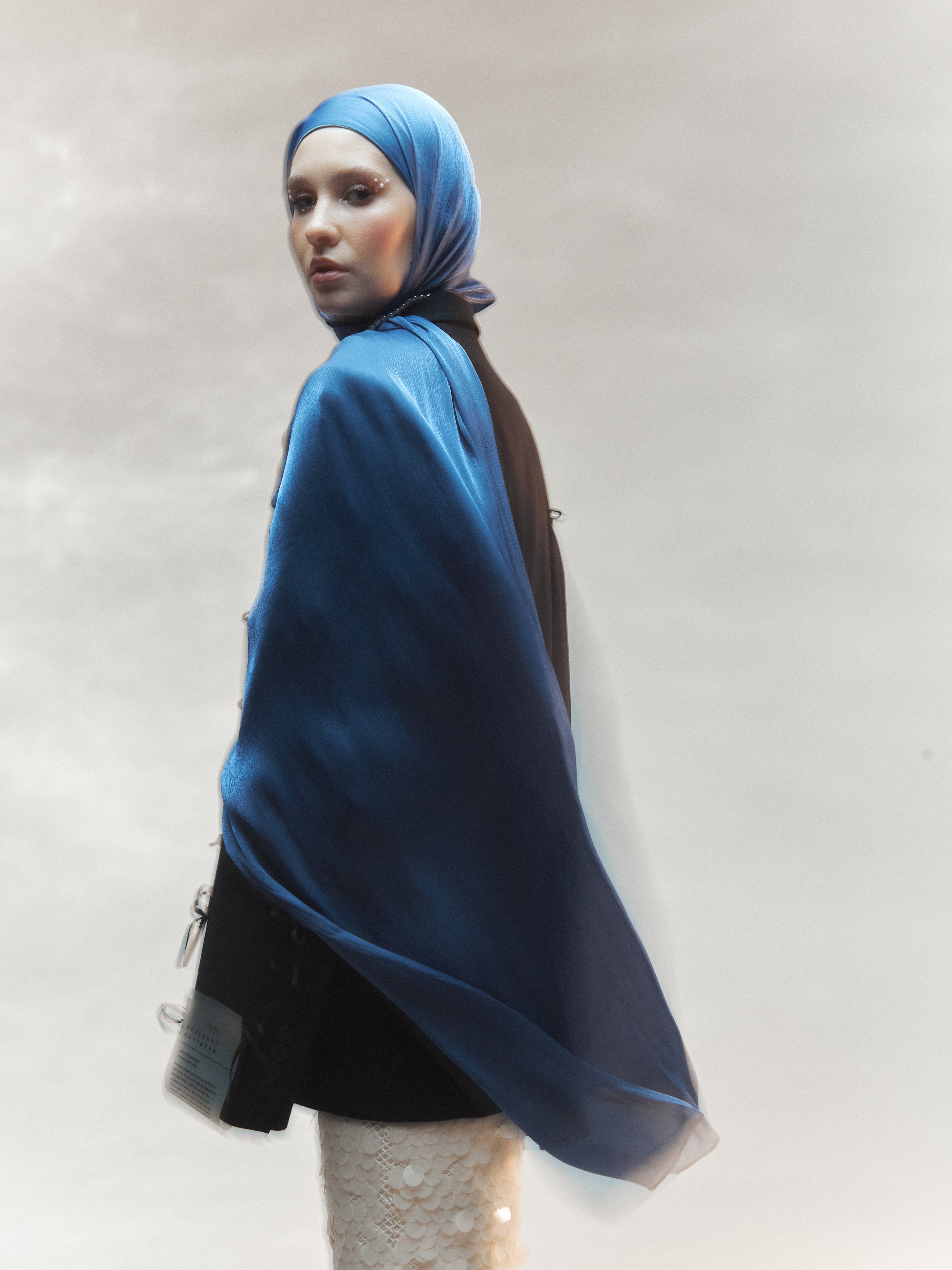 Woman wearing a steel blue ready-to-wear silk hijab styled with a black blazer and white sequin skirt, standing confidently against a soft, blurred backdrop.